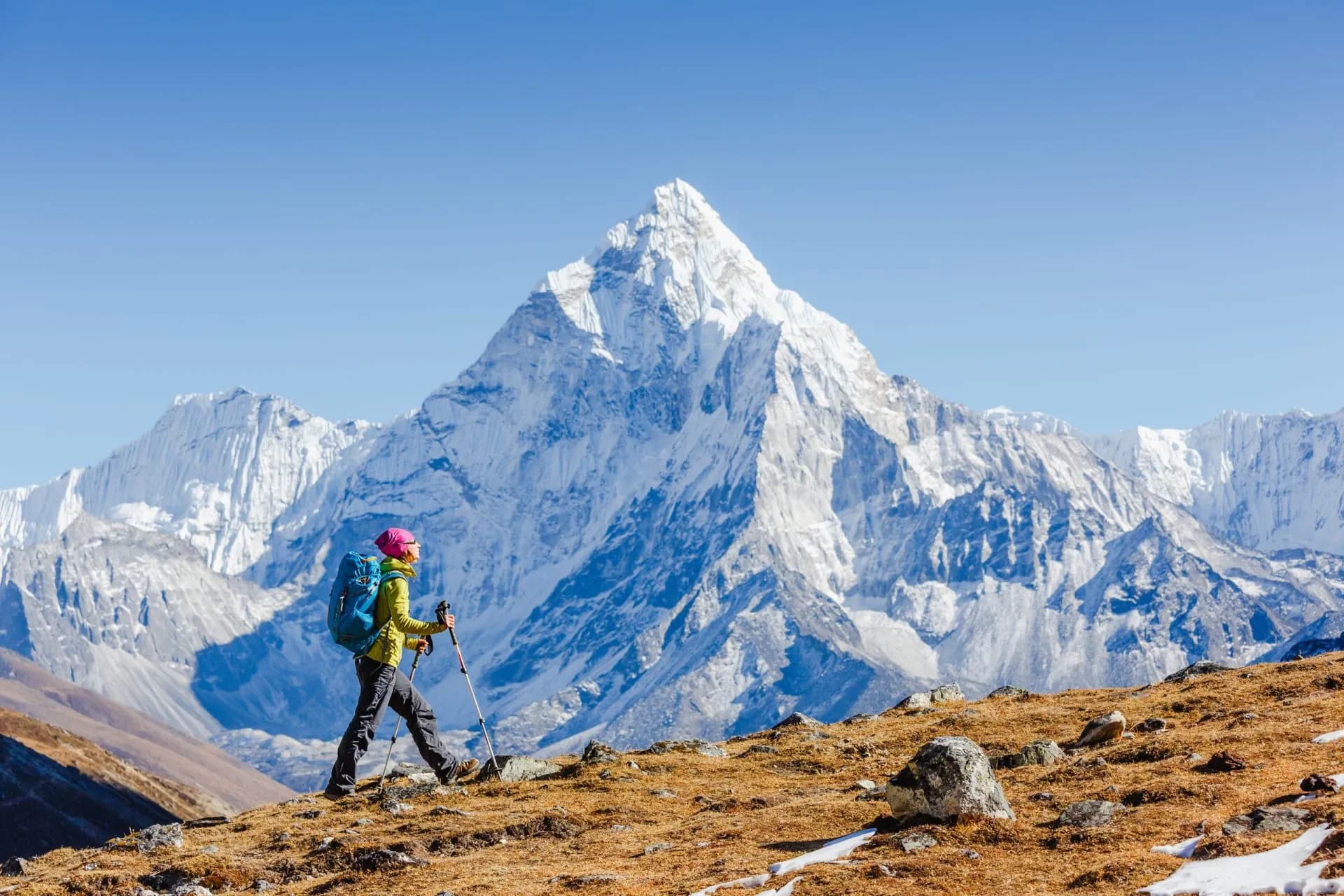 Hiker trekking with poles below massive snow-covered mountain peaks under a clear blue sky.
